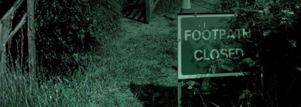 A green hued, low‑light outdoor scene showing a narrow grassy footpath surrounded by dense vegetation. On the right side of the image, a rectangular sign stands on metal legs with the words “FOOTPATH CLOSED” in large, reflective white letters. The sign is partially obscured by overgrown leaves and branches. Behind the sign, the path continues into a darker area, with an orange traffic cone placed near a wooden gate or barrier. 